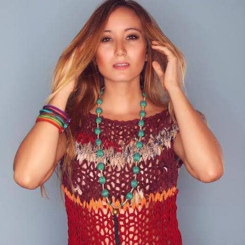 Woman wearing long red knit tunic top, turquoise jewelry, and standing in front of a gray wall