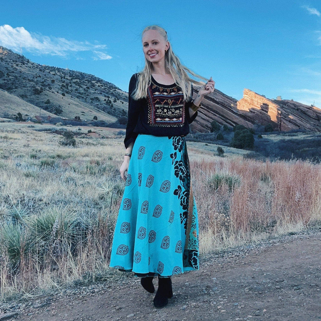Woman in embroidered black top and blue patterned skirt posing in a scenic landscape.