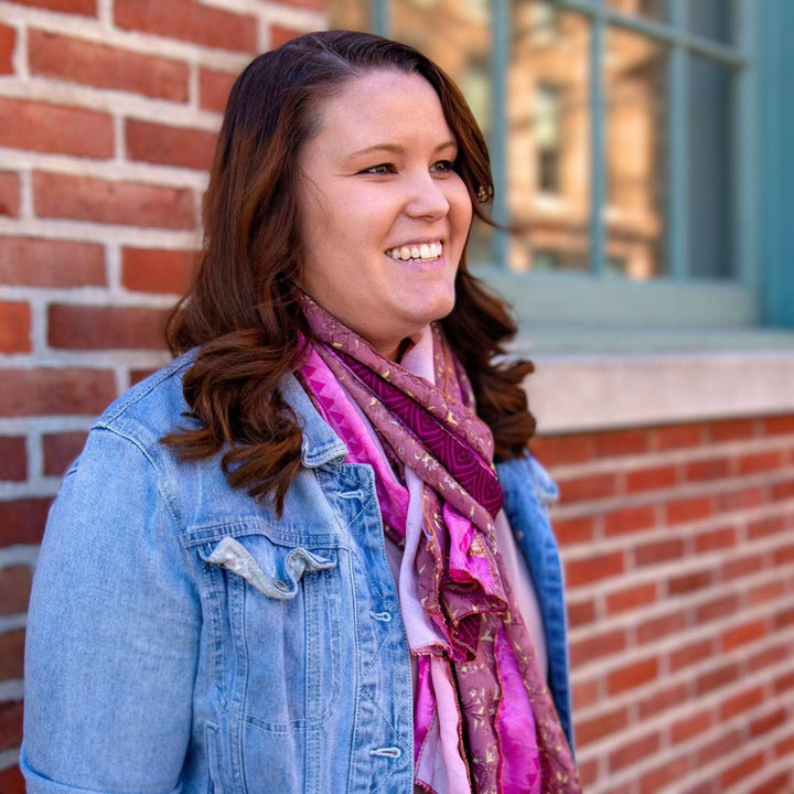 Woman smiling in denim jacket and purple scarf, leaning on brick wall.