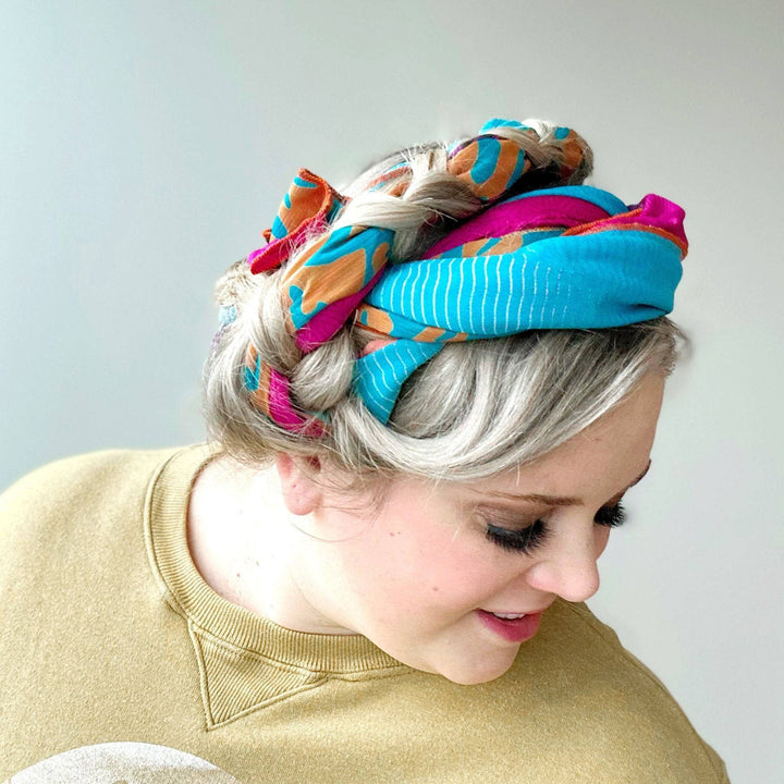 Woman with platinum hair wearing colorful scarf as headband. Yellow top, smiling, looking down.