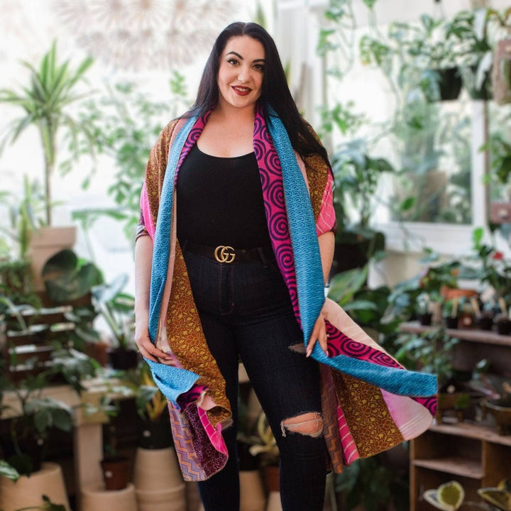 Woman in black outfit with colorful patchwork shawl. Plants in background. Smiling at camera.