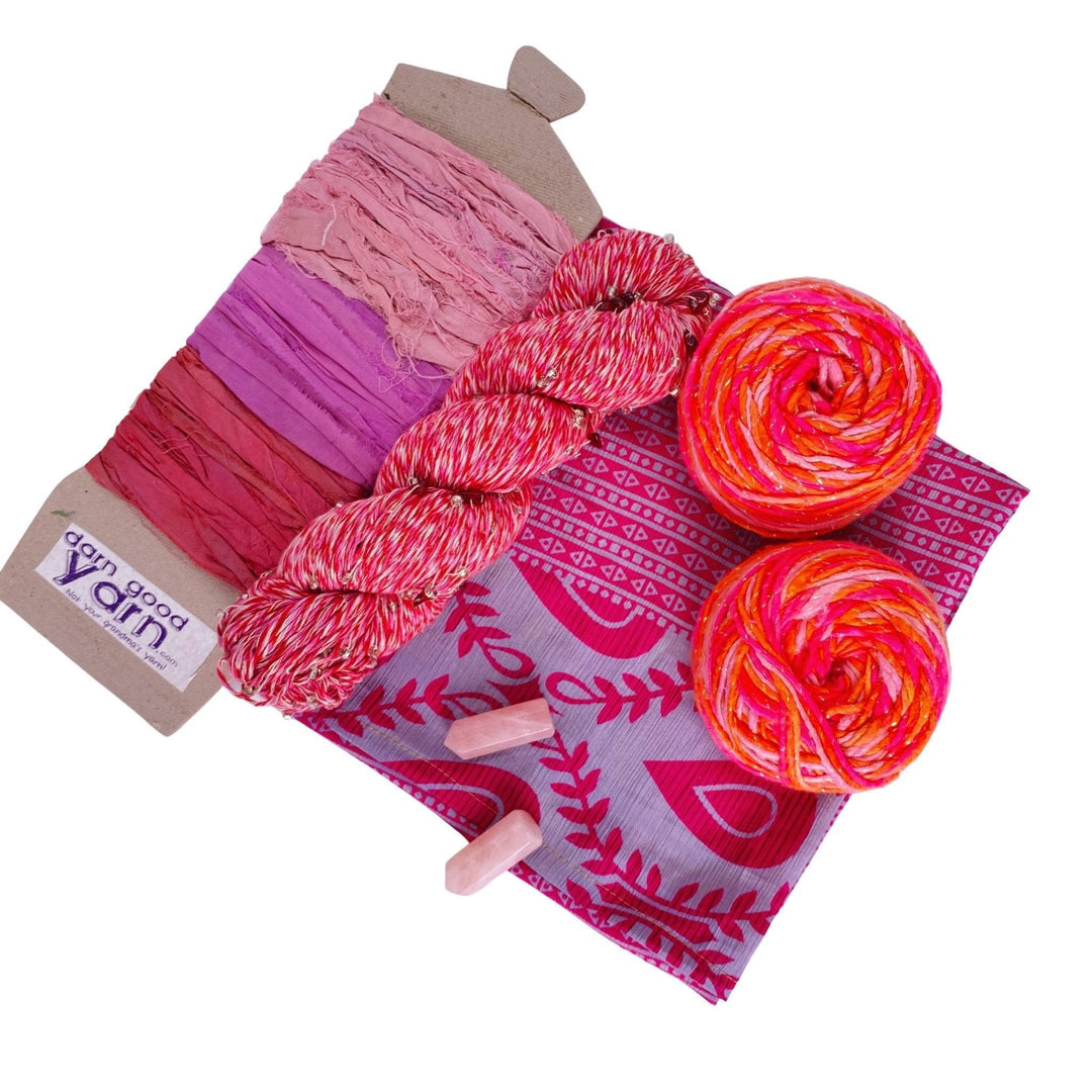 Rose Quartz energy bundle with all items showing in front of a white background. 2 rose quartz crystals, 2 skeins sparkle worsted weight silk, 1 skeins chakra beaded cotton yarn, 3 color sari silk ribbon sample card, and coordinating furoshiki.