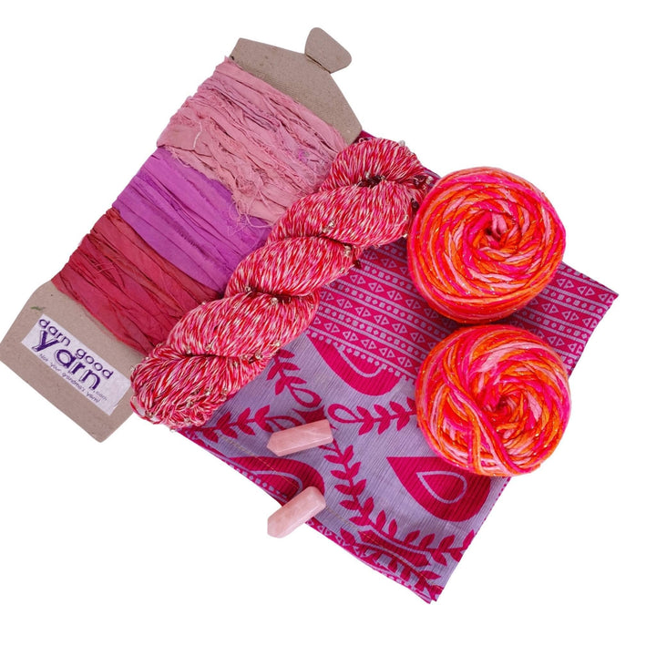 Rose Quartz energy bundle with all items showing in front of a white background. 2 rose quartz crystals, 2 skeins sparkle worsted weight silk, 1 skeins chakra beaded cotton yarn, 3 color sari silk ribbon sample card, and coordinating furoshiki.