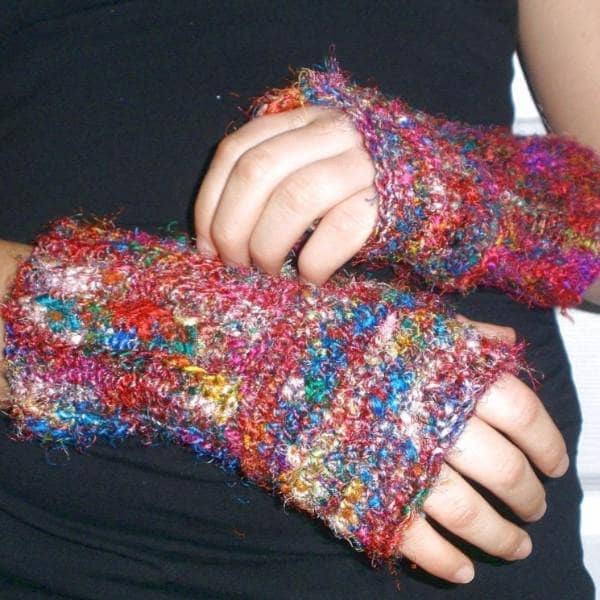 A person wearing colorful, textured fingerless gloves made of a fuzzy, multicolored yarn. The gloves cover the wrists and part of the hands, leaving the fingers exposed.
