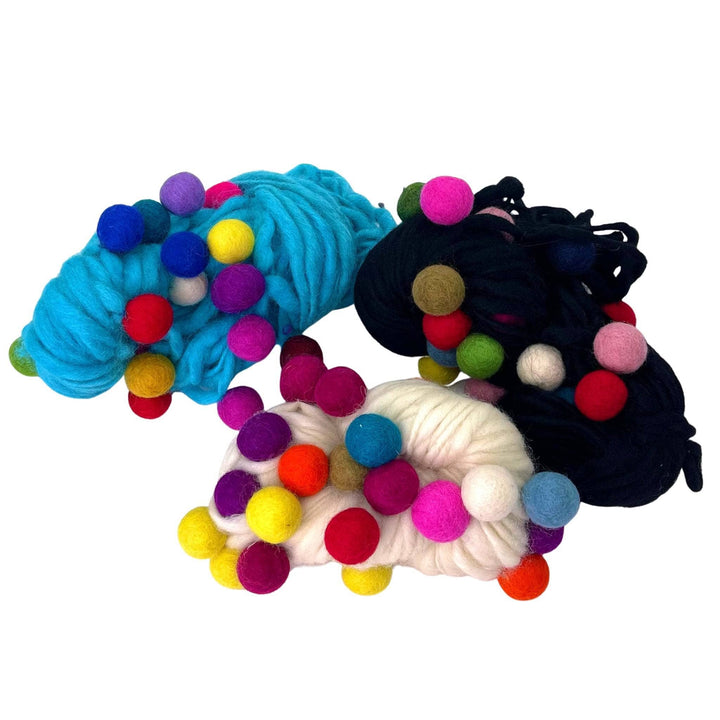 Three skeins of yarn with colorful felt balls, in blue, black, and white bases.