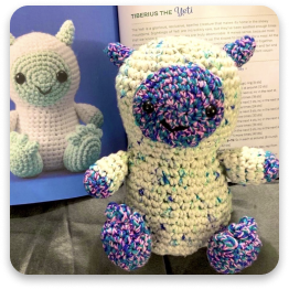 A crocheted stuffed animal, a blue and purple yeti, with a pattern book behind it.
