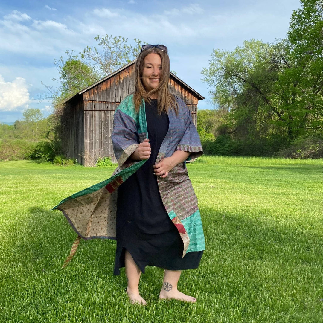 Model is outside in a field wearing a long kantha robe. In the background there is a barn. and blu skies. 