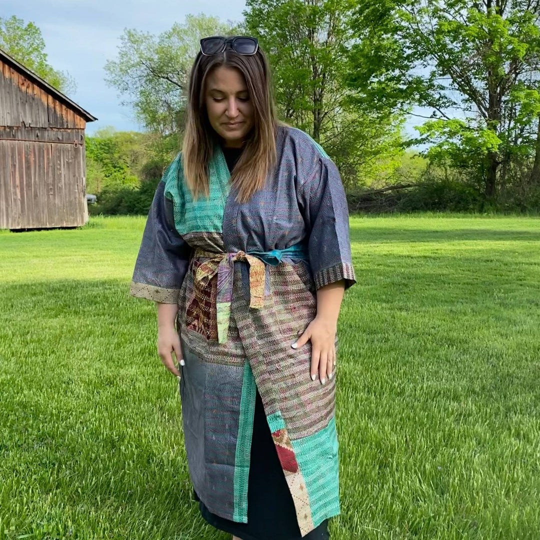 Model is standing outside in front of a barn wearing a one of a kind kantha jacket tied in the front . 