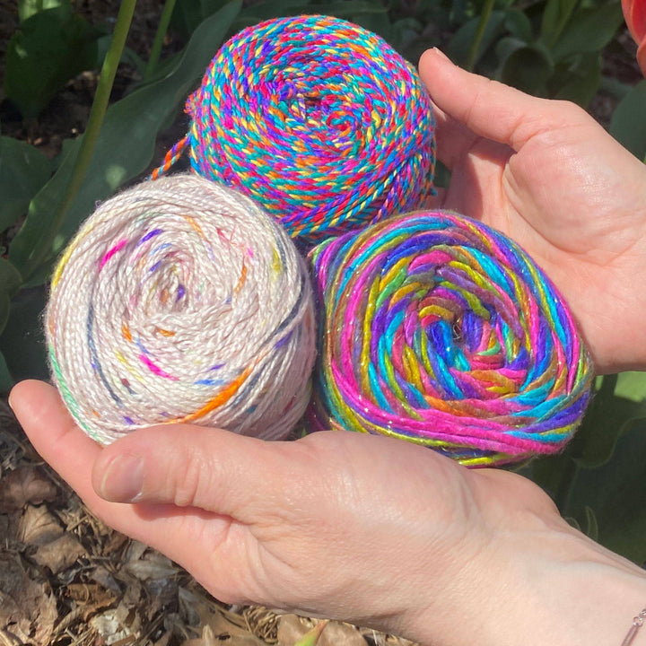 A hands holding three colorful yarn balls, featuring bright hues of pink, blue, and white