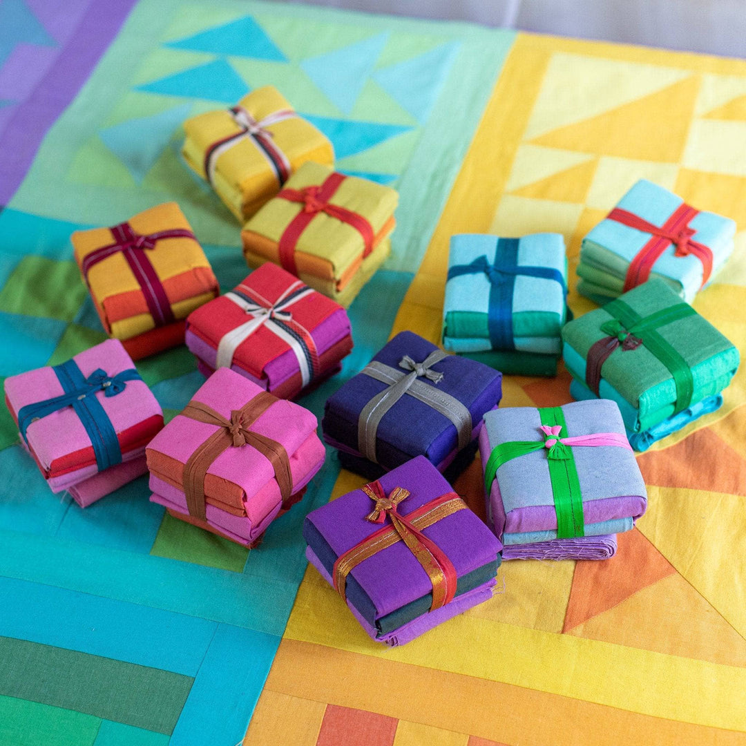 12 cotton fat quarter packs in assorted colors sitting on top of a colorful quilt.