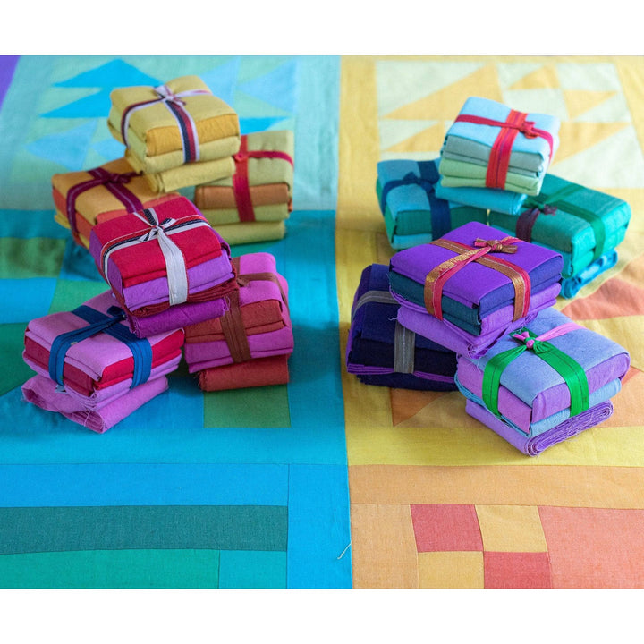 12 cotton fat quarter packs stacked in two piles (left and right) sitting on blocks of a completed rainbow quilt kit.
