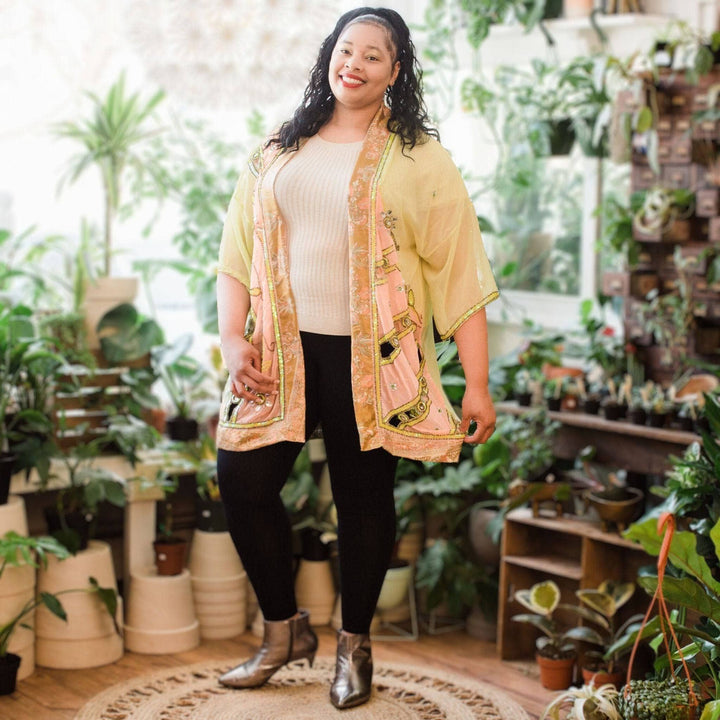 Model is wearing a light sheer yellow sophia silk duster while standing in front of potted plants. 