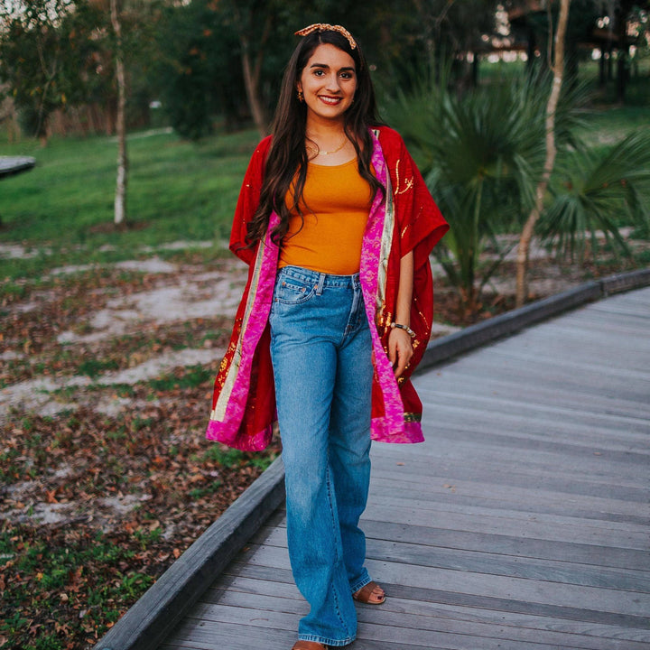 Woman in red shawl, orange top, and blue jeans stands on a wooden path with greenery.