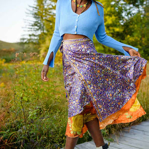 A woman wearing a purple and orange Sari Wrap Skirt walking on a boardwalk surrounded by fields of green in Spring-Like setting