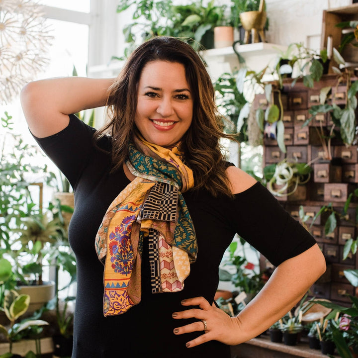 Model is wearing a one of a kind sari silk medley scarf. In the background there are some potted plants. 