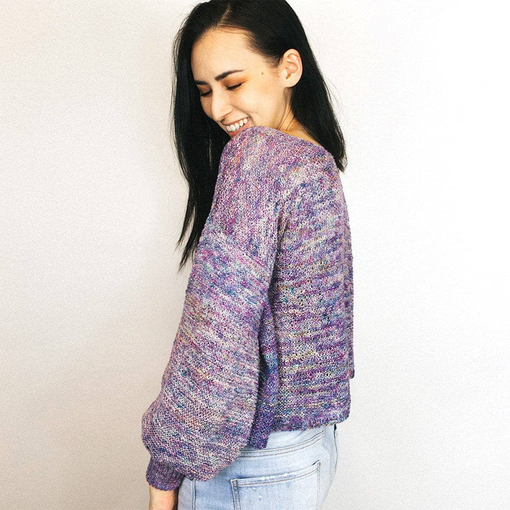 Woman wearing Zephyr Tides Cardi in Tidal Pool (purple) against a white wall