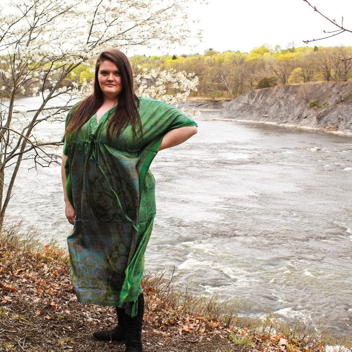 Model wearing sari silk maxi kaftan dress in green while standing outside with some budding trees and a river in the background.