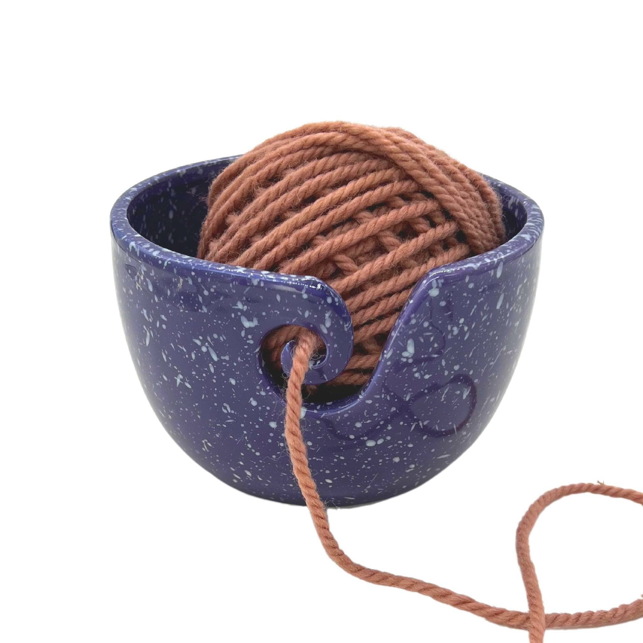 Purple with white speckled ceramic yarn bowl with salmon colored yarn inside of it on a white background 