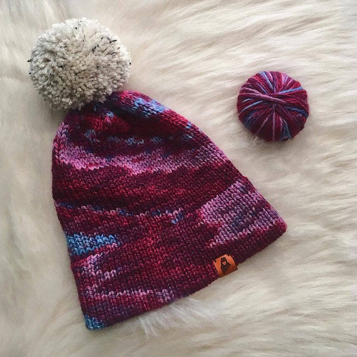 Purple knit beanie with large white pom pom next to purple yarn ball on a white fur surface