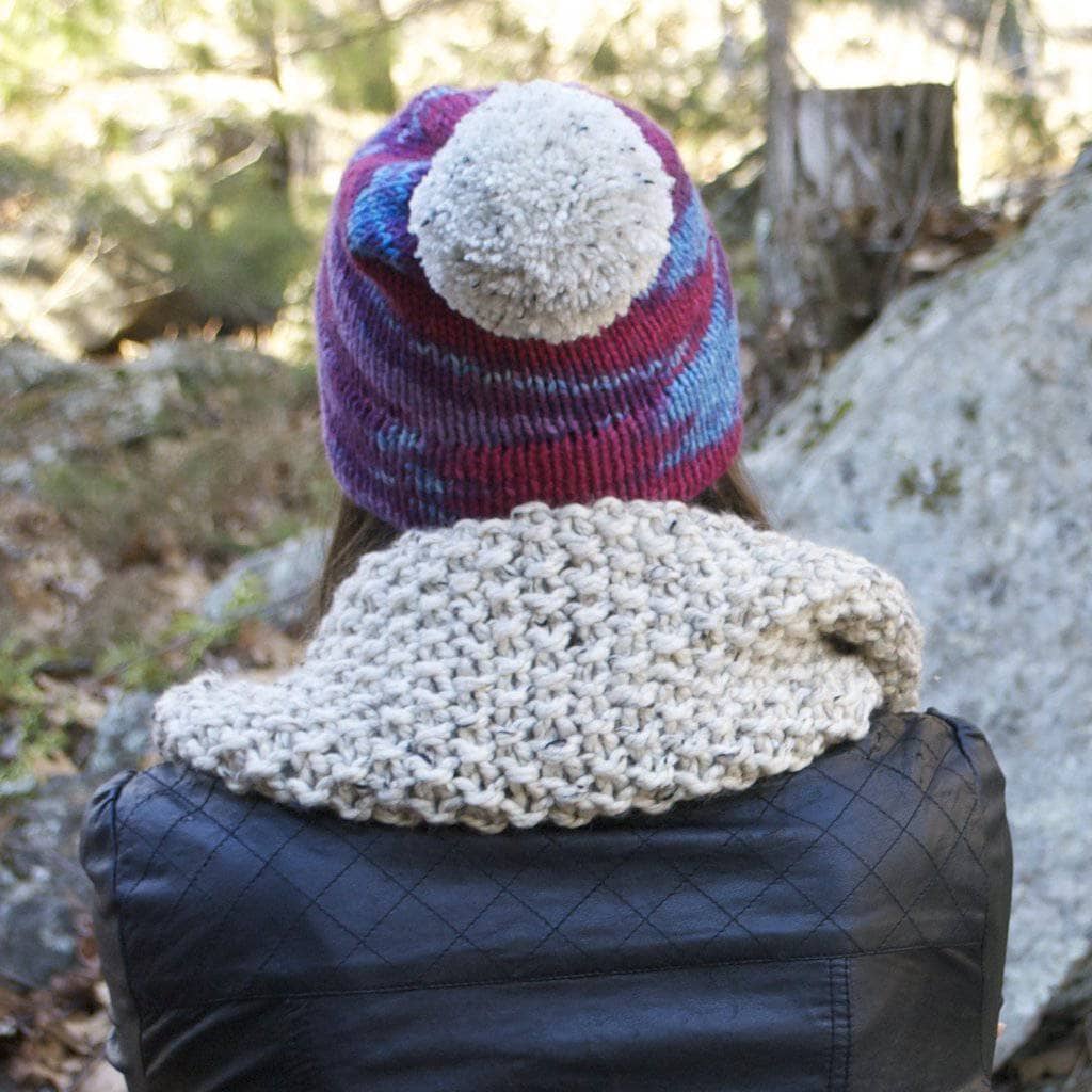 Woman wearing a purple beanie with white pom pom and white scarf and leather jacket facing a landscape background