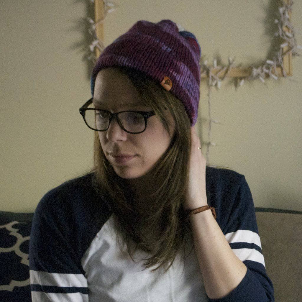 Woman wearing a purple beanie, glasses, and baseball tee sitting in front of a beige wall