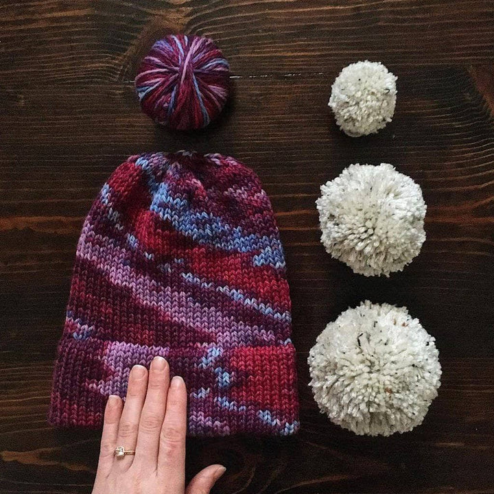 Purple beanie with white pom poms and purple yarn ball on a wooden surface