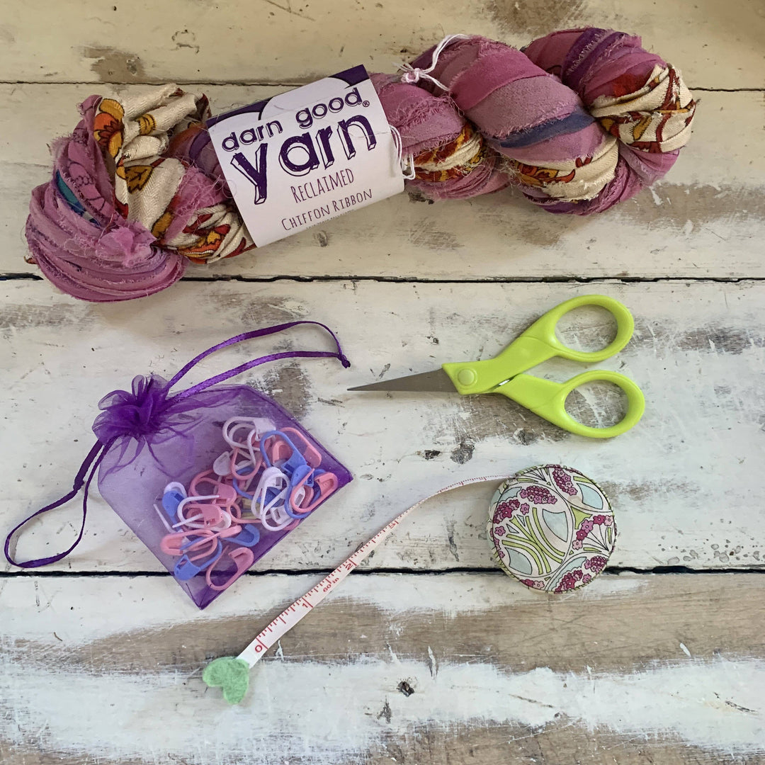 Tools and materials for finger knit infinity scarf in front of a wood background. Reclaimed chiffon ribbon yarn, multicolor pastel stitch markers, scissors, tape measure.