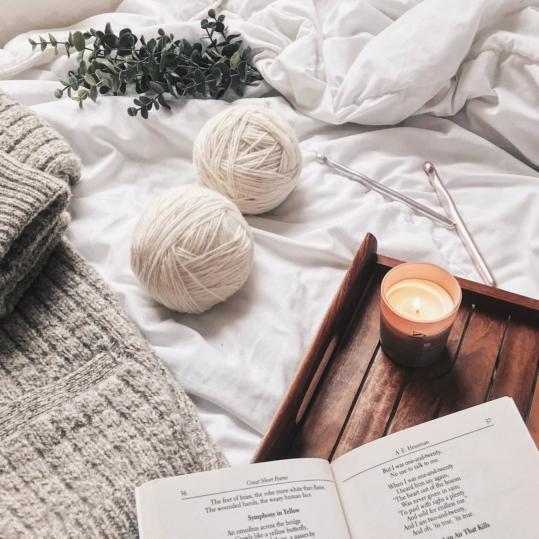 The Calla Cowl Bandana Crochet Kit next to a wooden tray with a book and candle on a white bed