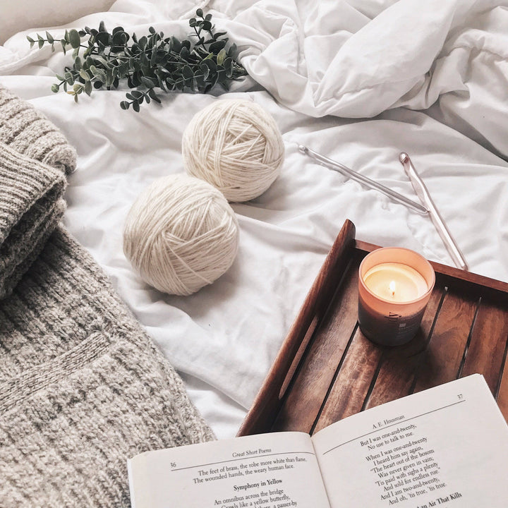 The Calla Cowl Bandana Crochet Kit next to a wooden tray with a book and candle on a white bed