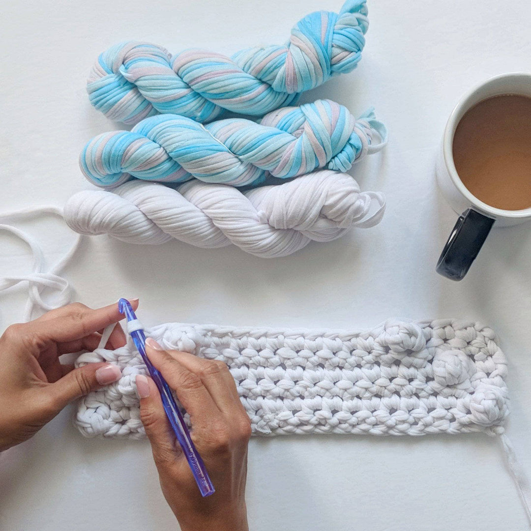 Partially made X Marks The Spot Wall Hanging with woman's hands holding a crochet hook, 3 skeins of yarn, and a cup of coffee