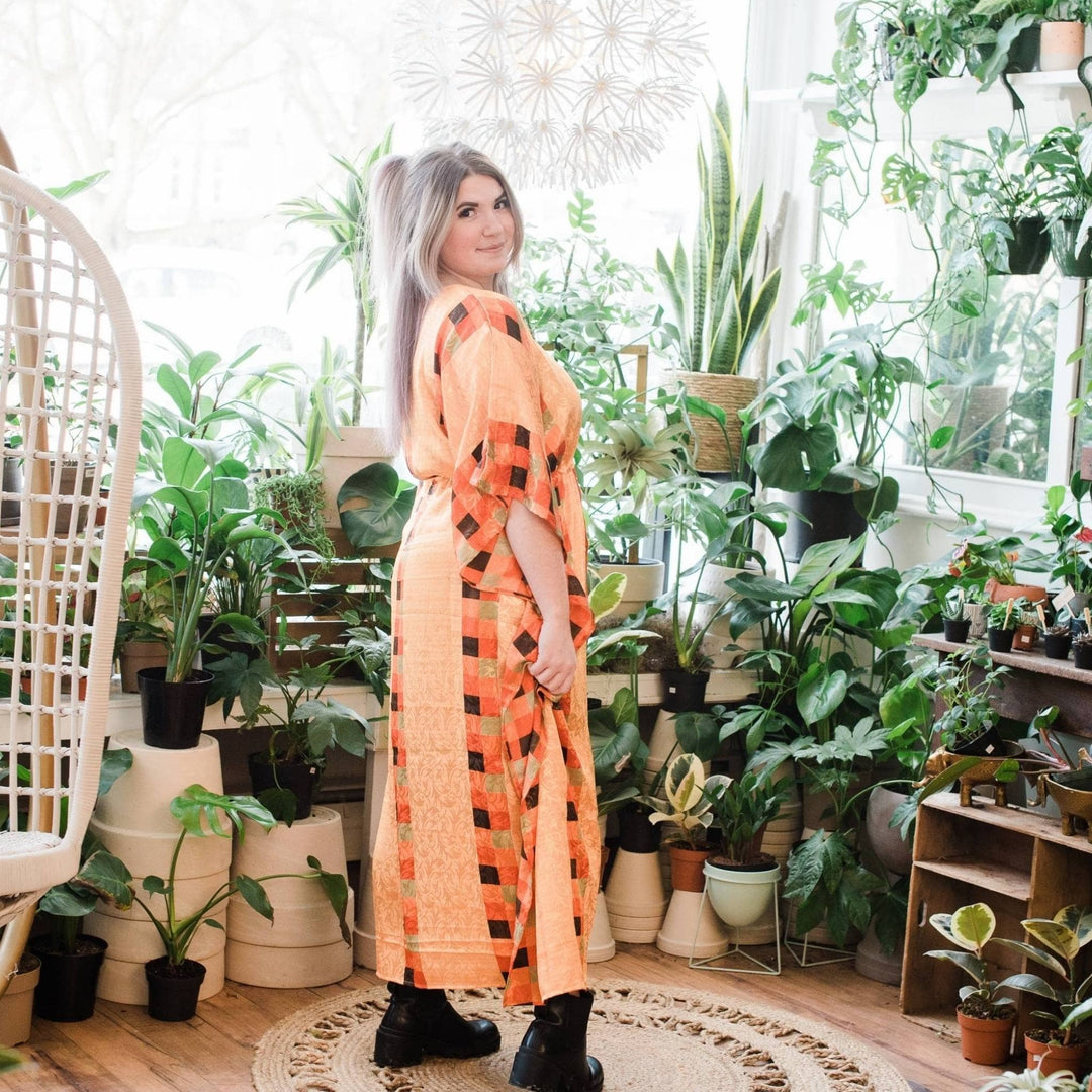 Model is turned to the side wearing an orange Aanya long kaftan while standing in front of green potted plants. 
