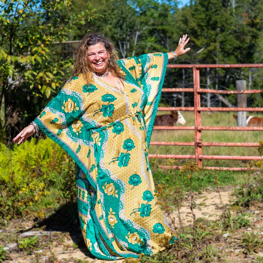 Model is standing in a field in front of a barn gate with cows in the background. Model is wearing a gold and green long aanya kaftan.