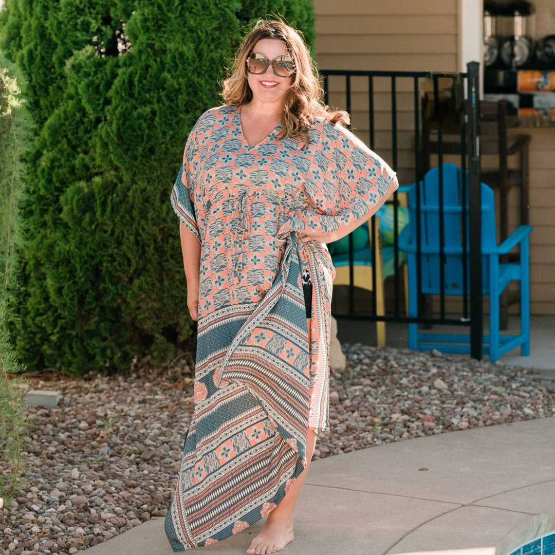 A woman standing by a pool wearing a sari adjustable floor length kaftan and big sunglasses. This kaftan is cream & teal colored with a paisley pattern all over.
