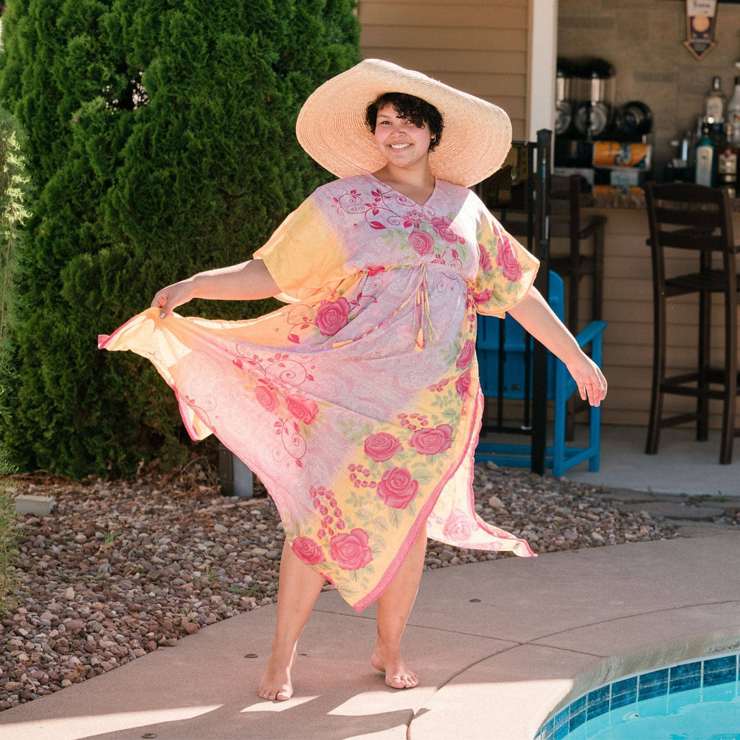 A woman standing by a pool wearing a long sari adjustable kaftan and a floppy sun hat. The kaftan is a light pink and cream with bright red roses all over it.