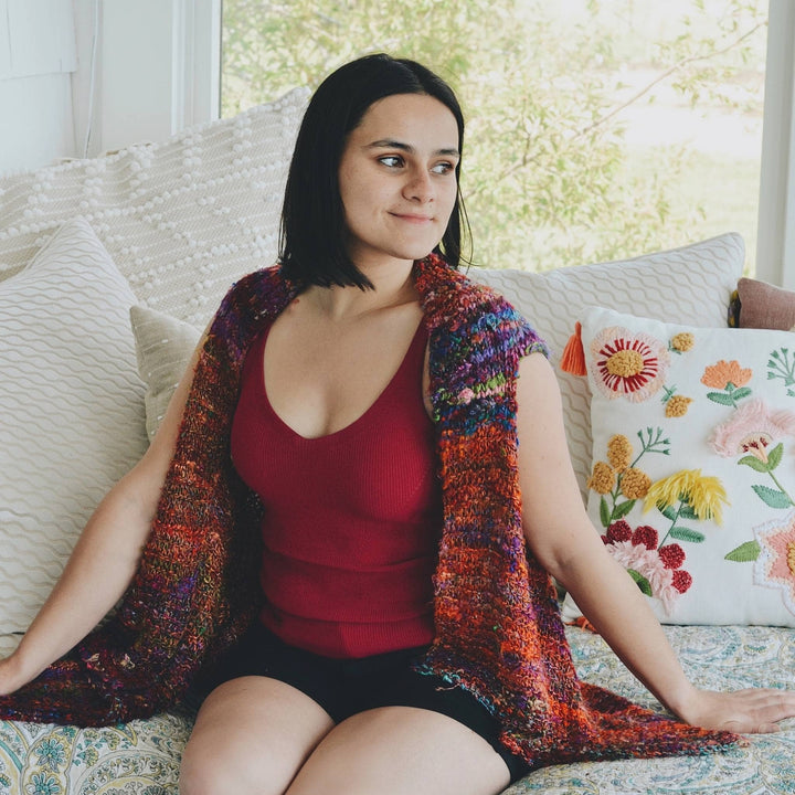 Model sitting on a couch wearing a knit adventure cardi in one of a kind kaleidoscope banana fiber yarn.