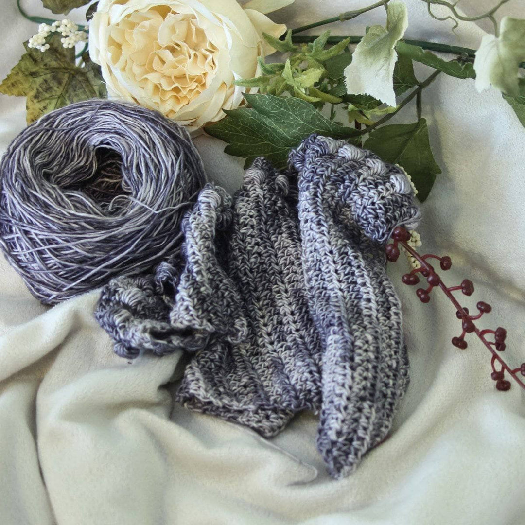 Andina Scarf in Date Night (grays)  partially-crocheted sitting next to yarn ball and flowers on a white background