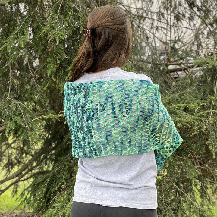 model wearing ariel shawl in variegated green colorway with greenery in the background.