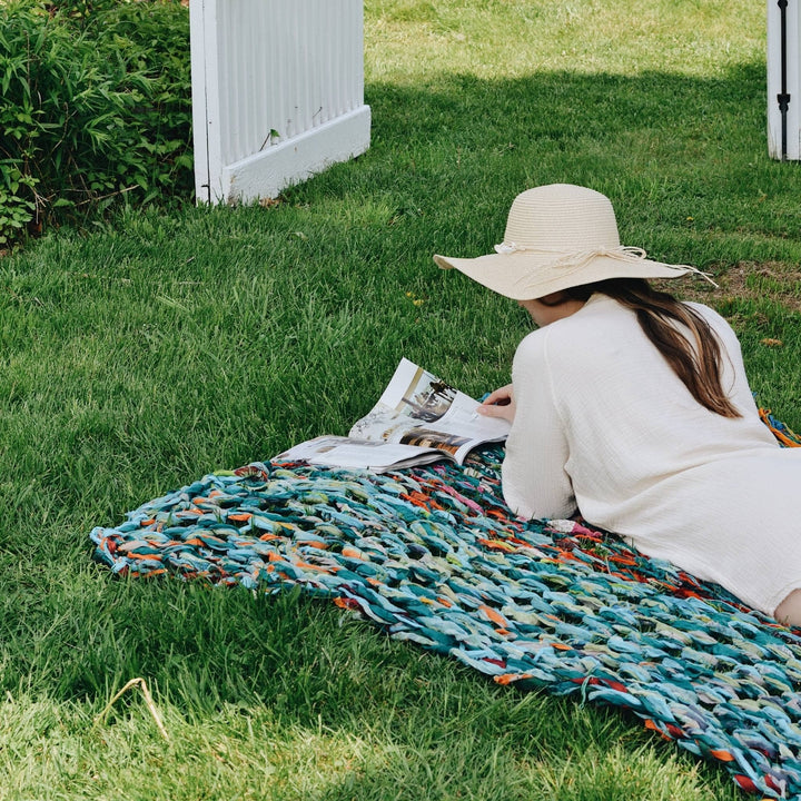Model relaxing on arm knit blanket out in the grass while reading a book with a white fence in the background.