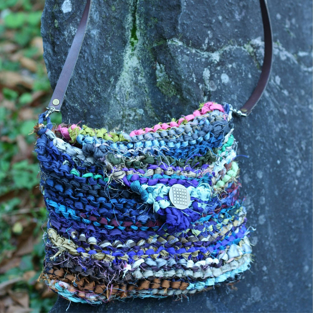 Knit around the world purse with silver button and leather strap with rock and greenery in the background.
