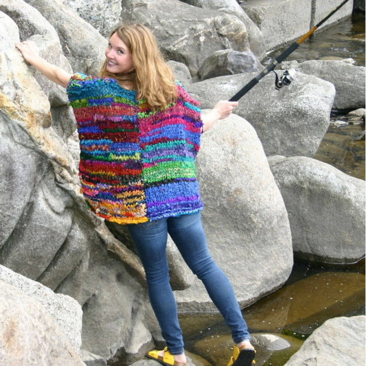 Founder Nicole Snow wearing artfully yours poncho while standing on and leaning against large grey boulders.