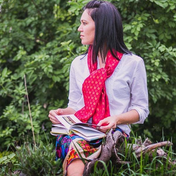 Woman wearing an Artisan Made Sari Silk & Cotton Scarf in red colors and a white shirt and sitting in front of greenery