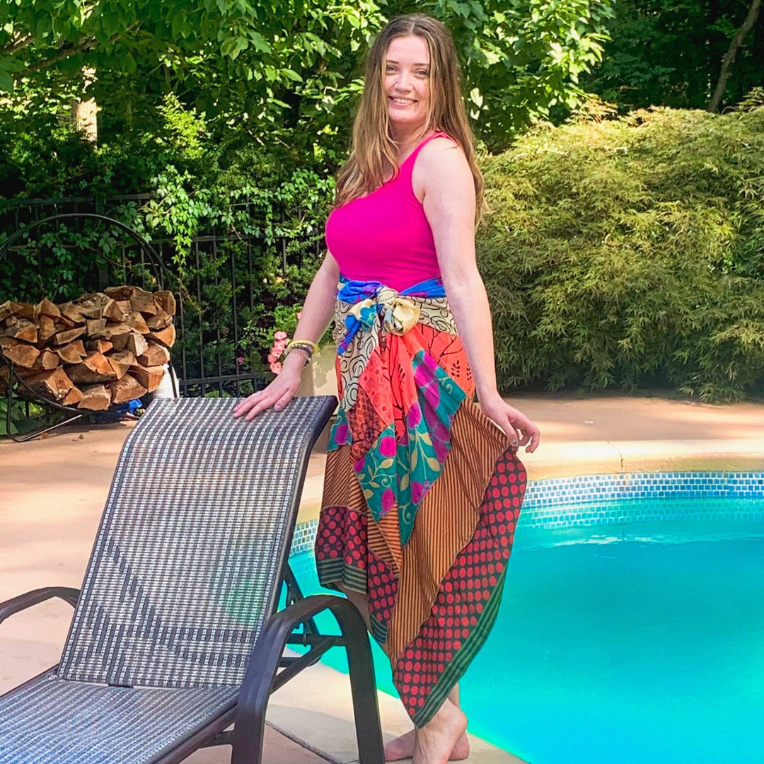Model is wearing an orange and red toned Bali Breeze Wrap as a baby suite coverup while standing next to a beach chair and a pool.