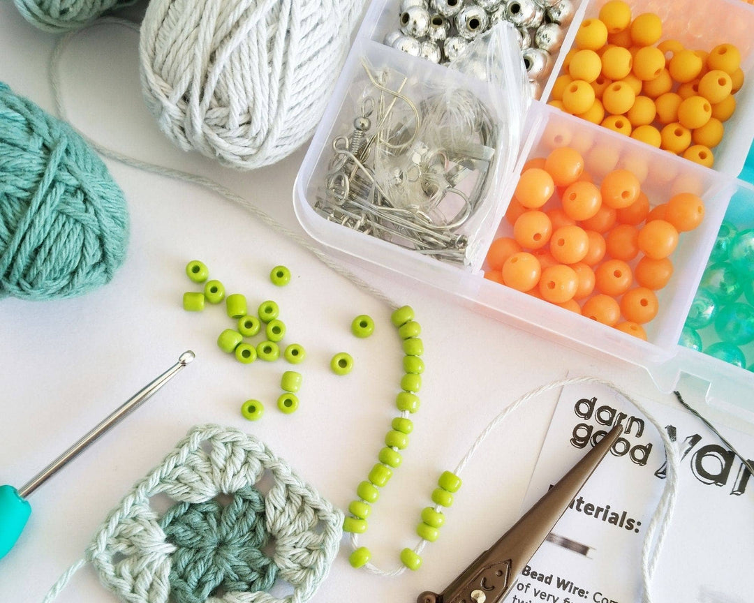 open bead case with beads, wires, and earring hooks showing in front of a white background. Yarn with beads strung onto it, a crochet hook, scissors, and instructions are scattered around.