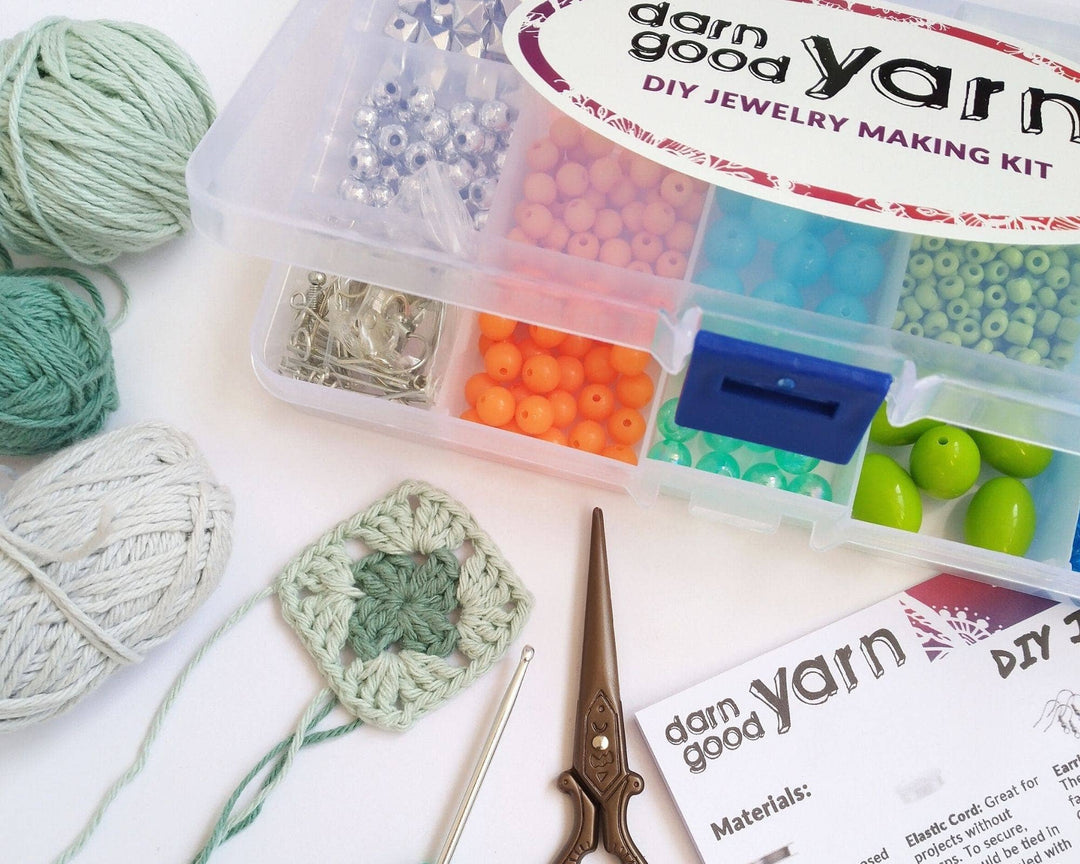 image of bead kit slightly opened with different yarn, crochet hook, scissors, and instructions scattered around, all in front of a white background.