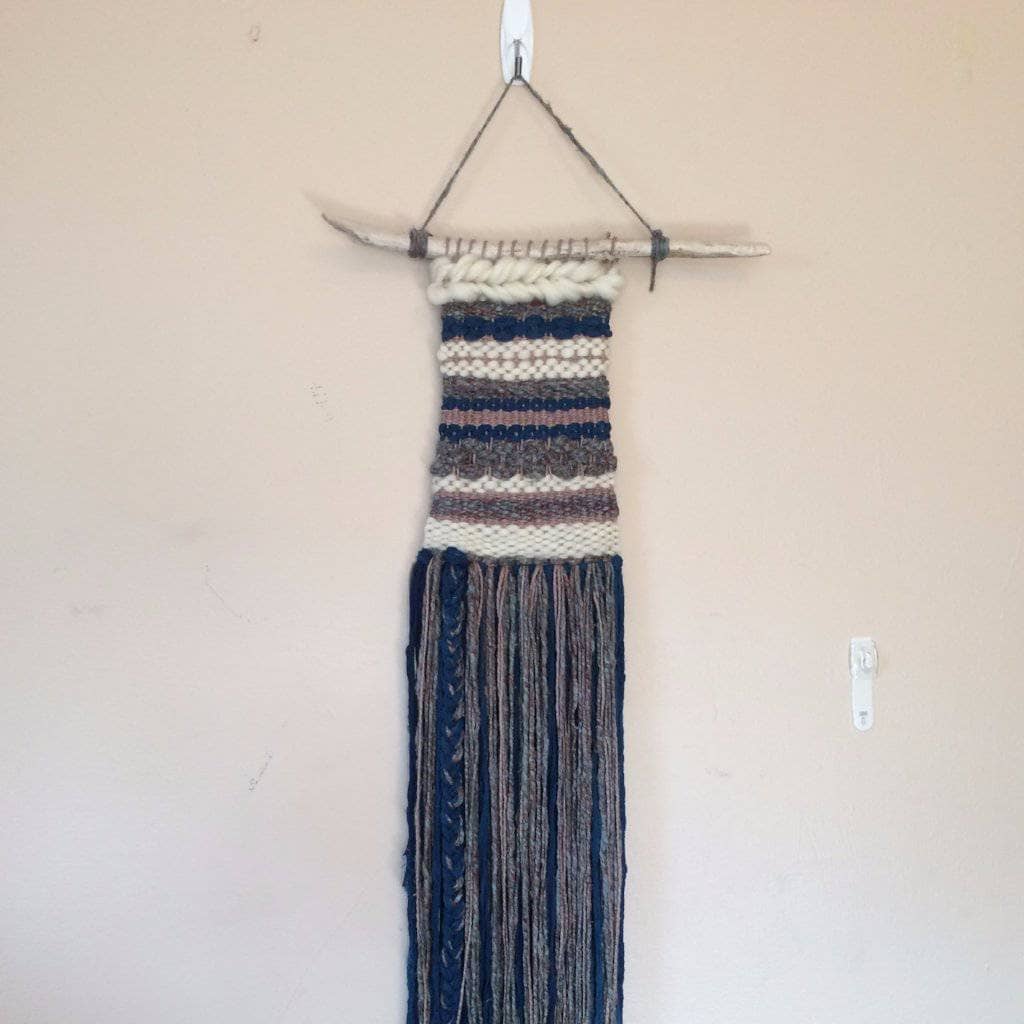 Woven tapestry in gray, navy, and white hanging from wooden stick on a white wall