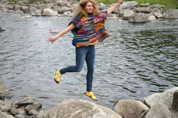 Woman wearing oversized wide-sleeve striped sweater jumping above a rock in front of a lake