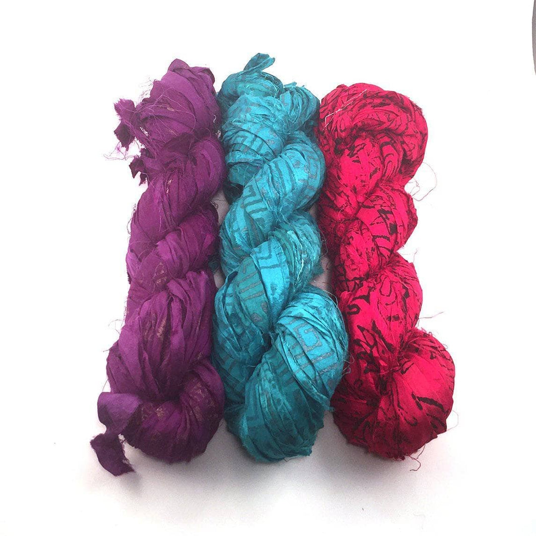 3 hanks of block printed sari silk ribbon yarn in front of a white background. Left to right Purple with gold, cerulean, and red paisley. 