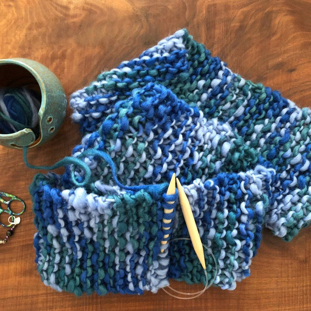 Blue Ridge Blanket Scarf laying on a wooden table next to a yarn bowl and wooden knitting needles