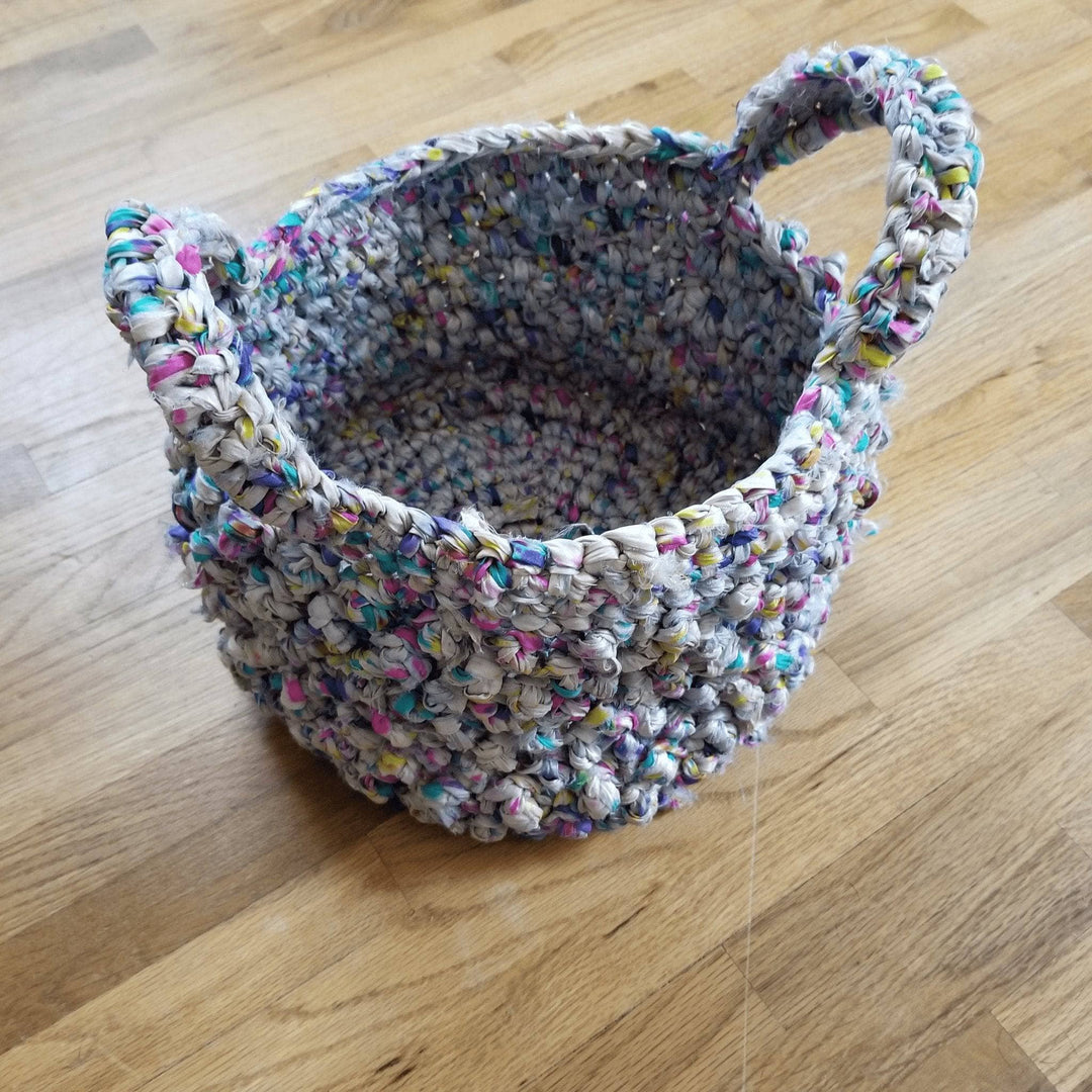 a white crochet basket with multicolored spots and handles sitting on the floor