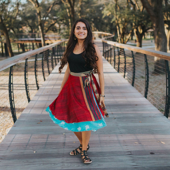 A model standing on a boardwalk wearing a Petit 00-02 Sari Wrap Skirt in Tea Length. The top layer is a rich red with gold accents and a wave pattern. The lover layer is a baby blue with white flowers across. She's paired the skirt with a plain tank top and matching gladiator sandals.